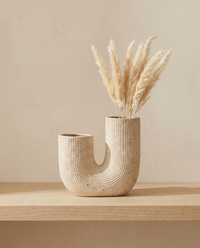Vase Arco mit Pampasgras in einer Oeffnung, Look auf Holzbrett vor warmer Wand